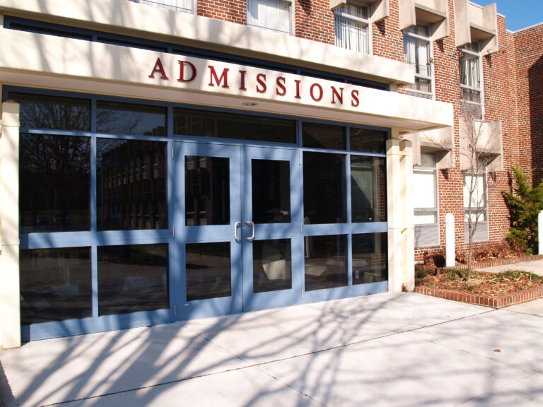 An exterior view of a college's Admissions office. The entrance features a set of clear glass double doors framed in a bold blue color. Above the doors, a sign reads 
