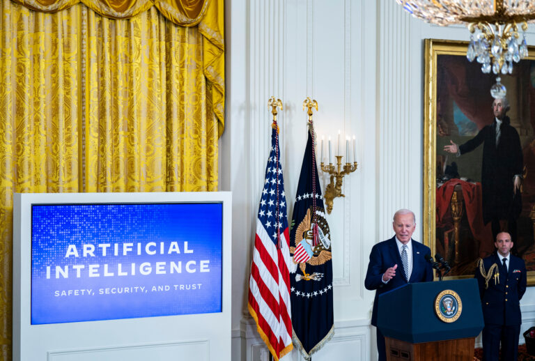 President Biden addressing the Executive Order on Artificial Intelligence, speaking at a podium with a digital screen displaying 